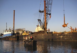Crane Lowering Concrete Into Water & Men Watching From Rowboat Near Long Island, NY